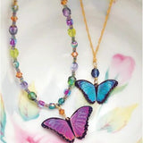 Discover the Green Flash Bella Butterfly Necklace by Holly Yashi: one features vibrant multicolored beads and a pink-purple butterfly, the other highlights a blue butterfly on a gold chain. Both are elegantly set against a floral backdrop.