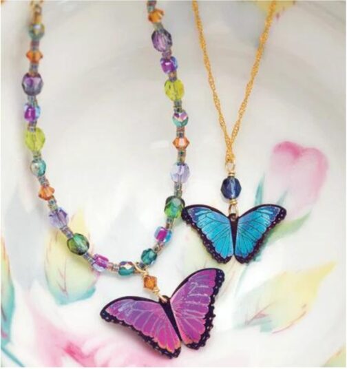 Discover the Green Flash Bella Butterfly Necklace by Holly Yashi: one features vibrant multicolored beads and a pink-purple butterfly, the other highlights a blue butterfly on a gold chain. Both are elegantly set against a floral backdrop.
