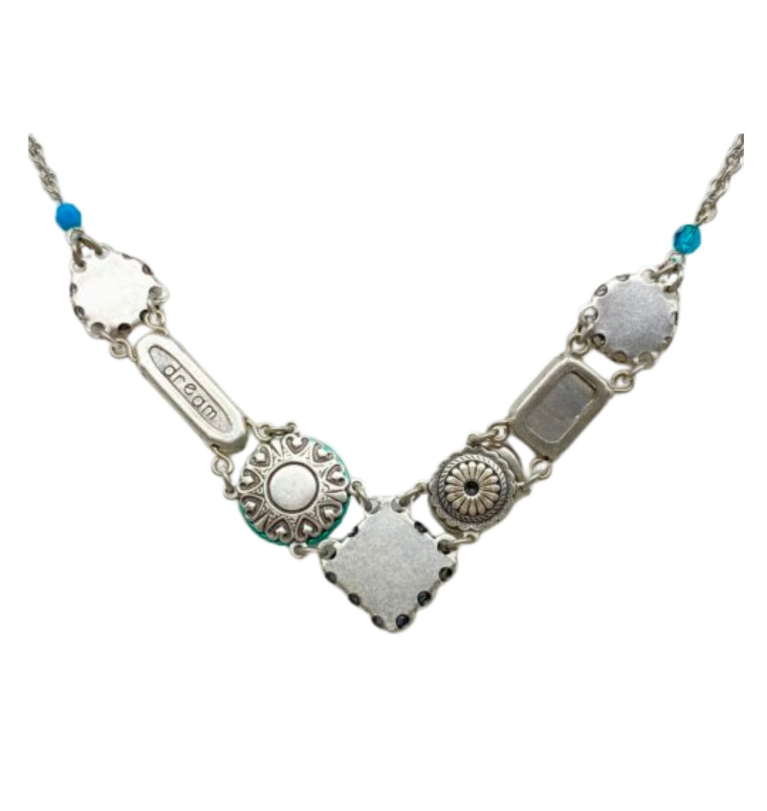 The La Dolce Vita V Multi-Color Necklace by Firefly is a handmade silver piece with a V-shaped decorative metal, engraved with a floral design and dream, featuring small blue beads, crafted through fair-trade practices.
