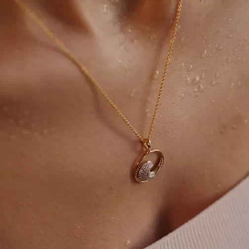 A close-up of the Gold Vermeil Ocean Wave Necklace by Shanore features a pendant with two intertwined rings—one textured, one smooth—resting on dewy skin and elegantly complementing a white top.