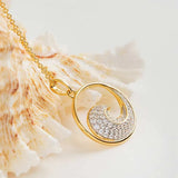 The Gold Vermeil Ocean Wave Necklace by Shanore, featuring a shell-inspired pendant with sparkling stones, is elegantly displayed next to a textured seashell on a light background.