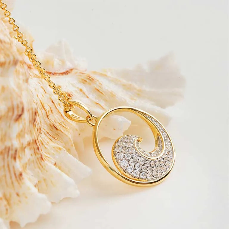 The Gold Vermeil Ocean Wave Necklace by Shanore, featuring a shell-inspired pendant with sparkling stones, is elegantly displayed next to a textured seashell on a light background.