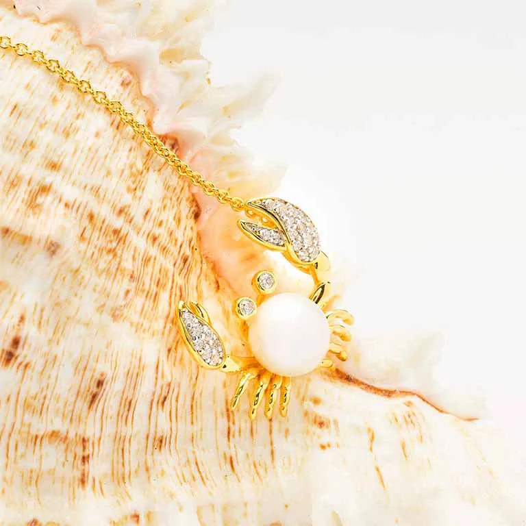 The Gold Vermeil Pearl Crab Necklace by Shanore features a crab-shaped pendant with white cubic zirconia, showcasing a pearl crab holding a round white pearl, set against a textured shell backdrop.