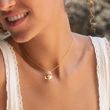 A person in a white sleeveless top smiles softly, with curly hair tied back. They wear Shanore’s Gold Vermeil Pearl Crab Necklace from the Ocean Jewelry collection, which elegantly catches the light against a beautifully blurred background.