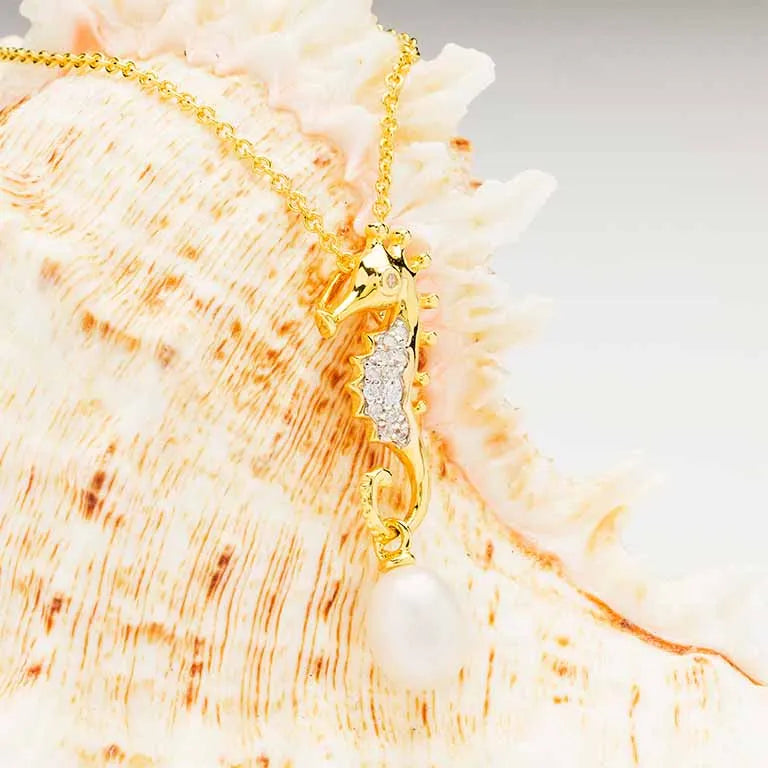 The Gold Vermeil Pearl Seahorse Necklace by Shanore features a seahorse pendant with crystals and a pearl, elegantly displayed on a textured seashell against a creamy white and peach gradient background.