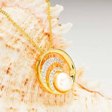 The Shanore Gold Vermeil Ocean Wave with Pearl Necklace is elegantly displayed against a textured seashell background, showcasing its curved gem-encrusted bands and large pearl pendant.