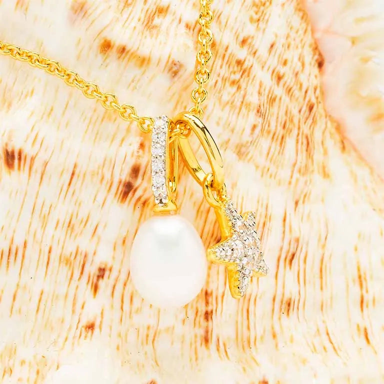 Shanores Gold Vermeil Starfish and Pearl Necklace - Ocean Jewelry features two pendants: a white pearl and a crystal-encrusted star. Set against a soft peach-toned textured backdrop, the delicate chain reflects the elegance typical of cubic zirconia jewelry.