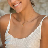 A person with curly hair and small hoop earrings smiles while wearing a light textured tank top. A Shanore Gold Vermeil Starfish and Pearl Necklace graces their neck. The softly blurred background highlights the serene moment.