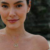 A person with long dark hair and bare shoulders smiles slightly, wearing the Gold Vermeil Blue Sapphire Compass Necklace - Ocean Jewelry by Shanore, paired with small earrings, against a blurred background.