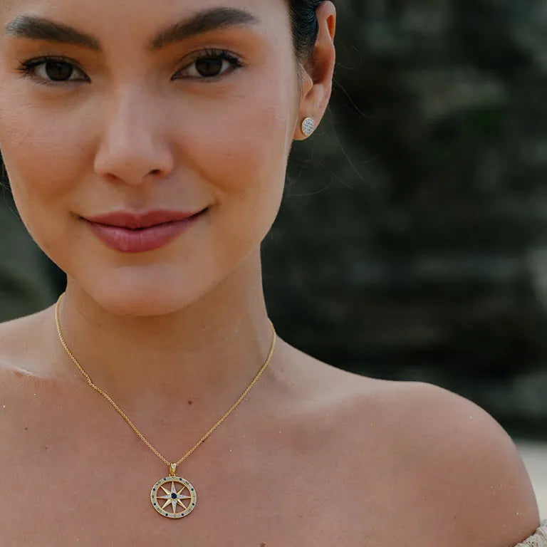 A person with long dark hair and bare shoulders smiles slightly, wearing the Gold Vermeil Blue Sapphire Compass Necklace - Ocean Jewelry by Shanore, paired with small earrings, against a blurred background.
