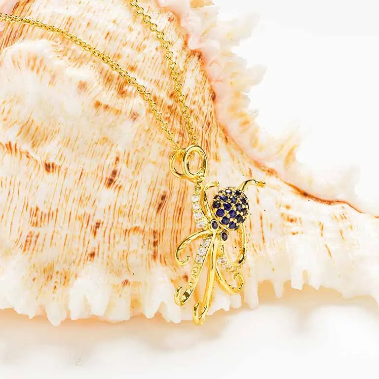 The Gold Vermeil Blue Sapphire Octopus Necklace by Shanore showcases an octopus pendant adorned with dark blue stones, including a striking blue lab-grown sapphire, set against a large seashell backdrop.