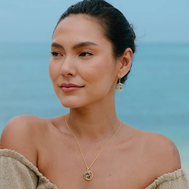 A dark-haired woman stands by the serene sea, wearing an off-shoulder beige top, and adorned with a Gold Vermeil Blue Sapphire Wave Necklace from Shanores Ocean Jewelry collection. She gazes slightly to the side, complementing her beach look with matching earrings.