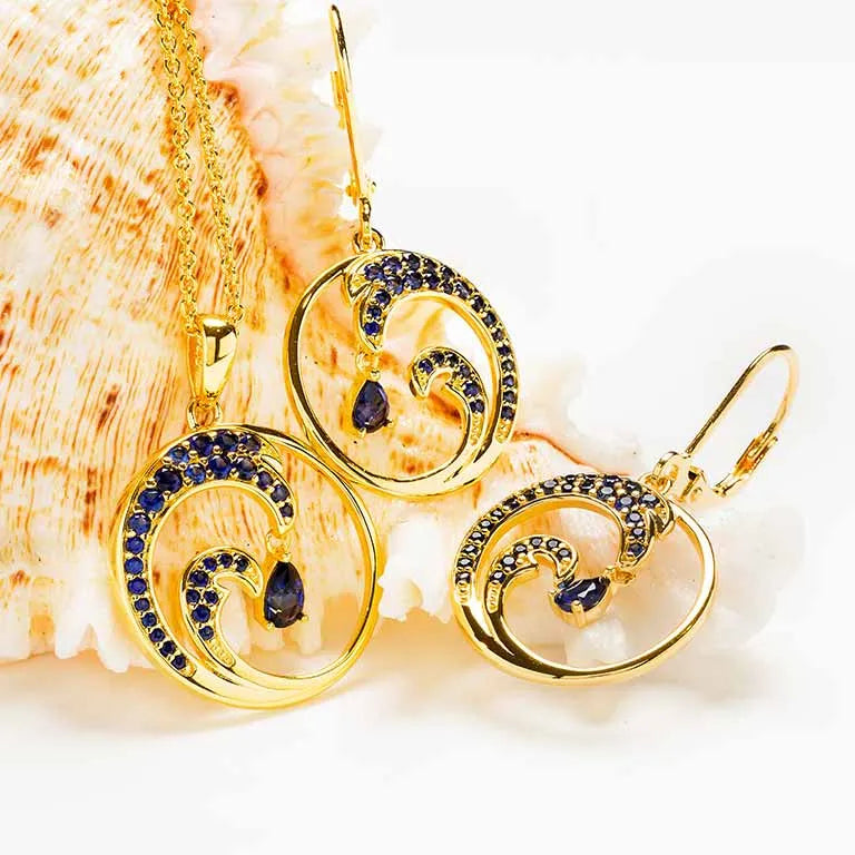 The Shanore Gold Vermeil Blue Sapphire Wave Necklace, part of the Ocean Jewelry collection, is showcased with intricate swirl designs and lab-grown sapphires, elegantly displayed on a light seashell background.