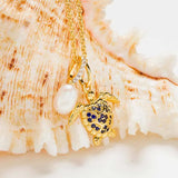 The Shanore Gold Vermeil Blue and White Sapphire Turtle Necklace gracefully hangs from a delicate chain, adorned with lab-grown sapphire and pearl charms, beautifully displayed against a textured seashell background.