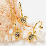 The Shanore Gold Vermeil Blue and White Sapphire Turtle Necklace, part of an ocean jewelry set with earrings, is adorned with lab-grown sapphires and elegantly placed on a large seashell. A small pearl charm complements the necklace.