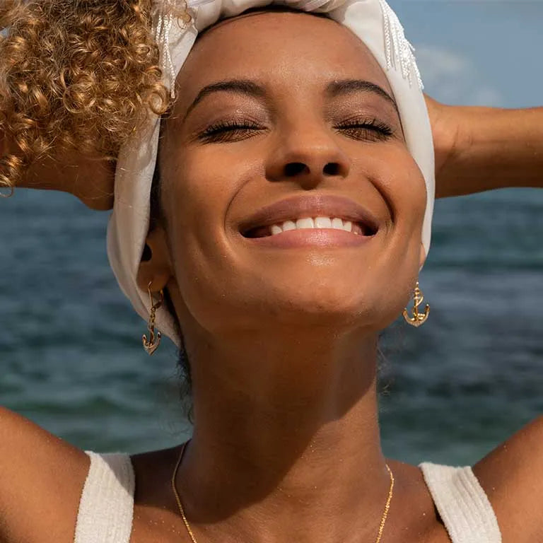 With a clear blue sky and ocean backdrop, a person with curly hair smiles joyfully, eyes closed, while wearing a white headscarf and Shanore’s Gold Vermeil Anchor Drop Earrings from the Ocean Jewelry collection, creating a sunny, tropical atmosphere.