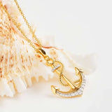 The Shanore Gold Vermeil Anchor Necklace - Ocean Jewelry features a shimmering cubic zirconia-adorned pendant on a delicate chain, resting against a textured seashell background, highlighting its nautical design and craftsmanship.