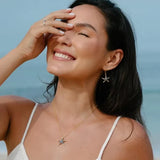 A woman with long dark hair smiles broadly on the beach, wearing a white spaghetti strap top. She showcases Shanores Gold Vermeil Blue Sapphire Starfish Necklace - Ocean Jewelry. Her hand gently touches her forehead as she gazes upwards, with the ocean glistening behind her.