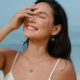A joyful woman with long dark hair touches her forehead, wearing 14k Gold Vermeil Blue Sapphire Starfish Earrings by Shanore. She stands against an ocean backdrop, dressed in a white spaghetti strap top.