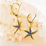 The Gold Vermeil Blue Sapphire Starfish Earrings- Ocean Jewelry by Shanore, featuring lab-grown sapphires, are elegantly displayed in front of a seashell.