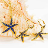 The Gold Vermeil Blue Sapphire Starfish Earrings by Shanore are displayed in front of a large seashell, featuring intricate detailing for an elegant, ocean-inspired design.