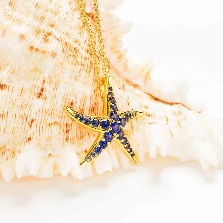 The Shanore Gold Vermeil Blue Sapphire Starfish Necklace features sparkling blue lab-grown sapphires, complemented by a delicate gold chain, and is elegantly showcased on a large textured seashell against a soft white background.