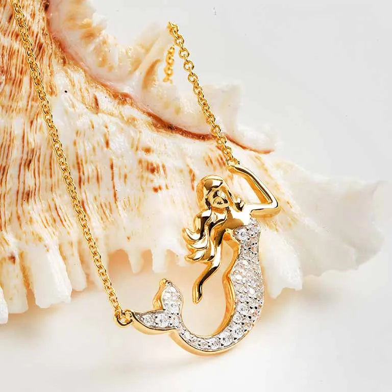 The Gold Vermeil Mermaid Necklace by Shanore, under the Ocean Jewelry collection, is showcased against a large seashell. It features an intricately detailed mermaid pendant embellished with shimmering cubic zirconia stones for an elegant touch.