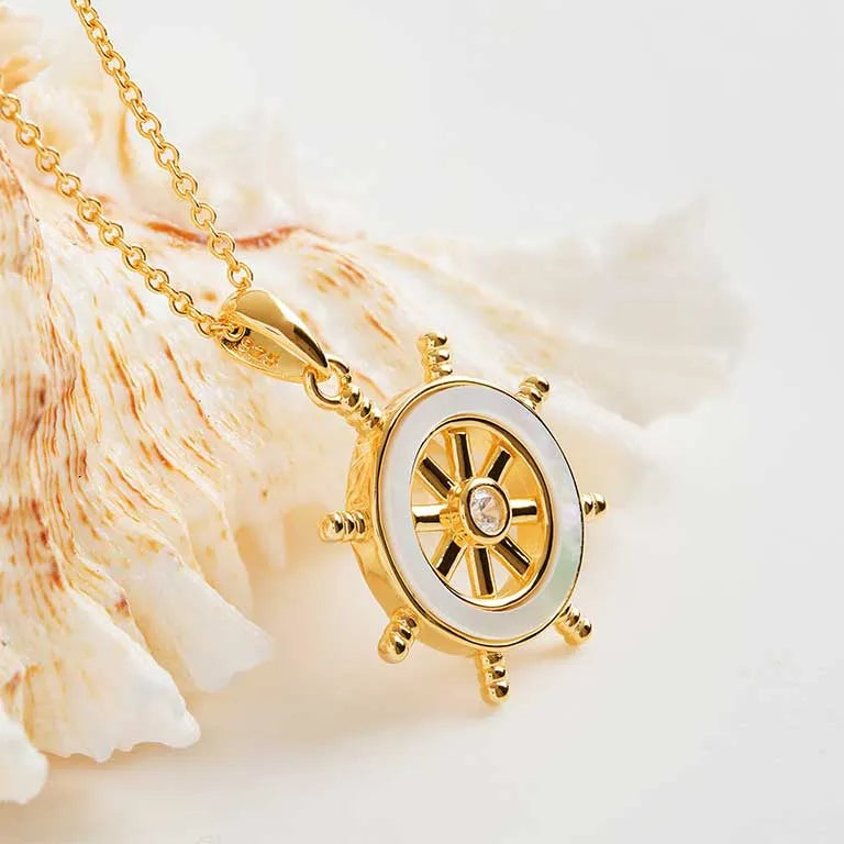 The Shanore Gold Vermeil Mother of Pearl Ship Wheel Necklace, an ocean jewelry piece, elegantly highlights a gem-centered ships wheel with white enamel on a textured natural shell backdrop.