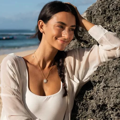 A woman with long dark braided hair leans against a rock at the beach, wearing a white top and sheer blouse. Around her neck glistens the Shanore Gold Vermeil Mother of Pearl Sun Necklace - Ocean Jewelry. The ocean sparkles under a clear blue sky in the background.