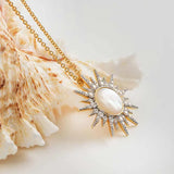 The Gold Vermeil Mother of Pearl Sun Necklace by Shanore features a mother of pearl center framed by cubic zirconia on a gold chain, elegantly displayed against a textured seashell backdrop.