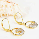 The Gold Vermeil Ocean Wave Earrings by Shanore feature a spiral design adorned with sparkling cubic zirconia, set against a soft white background with a textured seashell.
