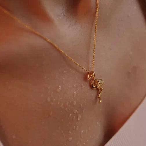A close-up captures the elegance of Shanores Gold Vermeil Pink Flamingo Necklace. Water droplets glisten on its surface, enhancing the pieces charm, while the warm and soft atmosphere accentuates its beauty.