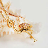 The Shanore Gold Vermeil Pink Flamingo Necklace features a flamingo-shaped pendant adorned with pink and orange gemstones. Its cubic zirconia wings dazzle on a delicate chain, set against a white seashell on a light background.
