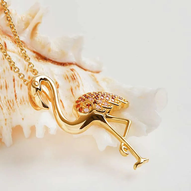 The Shanore Gold Vermeil Pink Flamingo Necklace features a flamingo-shaped pendant adorned with pink and orange gemstones. Its cubic zirconia wings dazzle on a delicate chain, set against a white seashell on a light background.