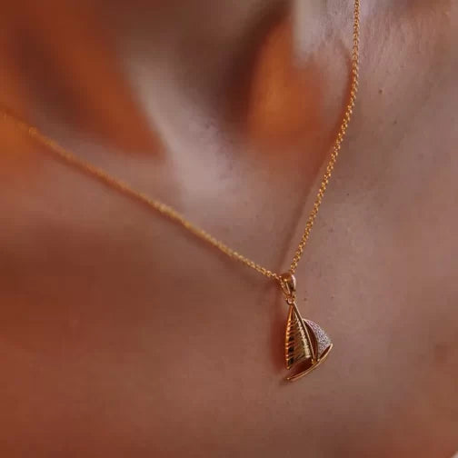 Someone is wearing the Shanore Gold Vermeil Sailboat Necklace - Ocean Jewelry, featuring a sailboat-shaped pendant. The softly blurred background highlights the necklace against their skin.