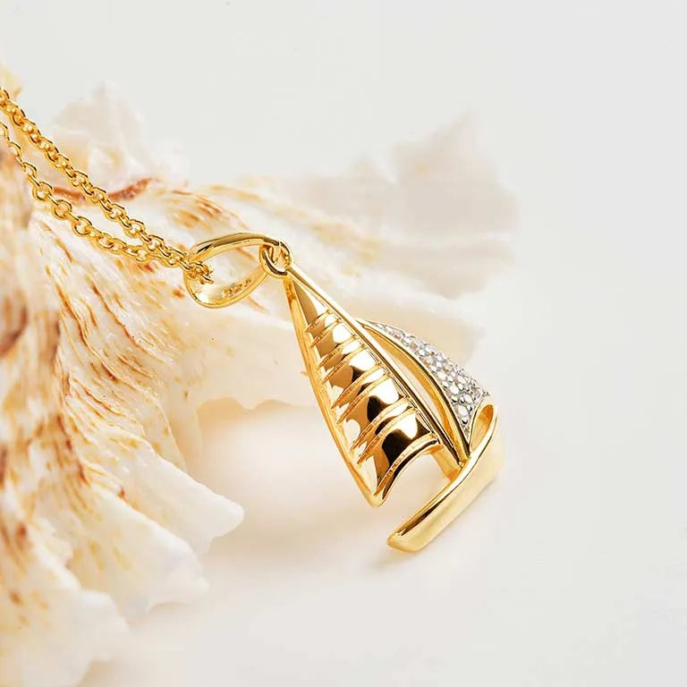 The Gold Vermeil Sailboat Necklace by Shanore, featuring a cubic zirconia-adorned sail, elegantly rests beside a textured seashell on a light background.