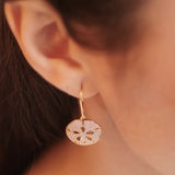 A womans ear adorned with Shanores Gold Vermeil Sand Dollar Earrings from the Ocean Jewelry collection, featuring a gold hoop with a circular pendant, delicately designed with cut-out shapes; the background is softly blurred.