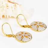 Gold Vermeil Sand Dollar Earrings from Shanore, featuring sparkling cubic zirconia, rest on a white surface with a seashell in the background.