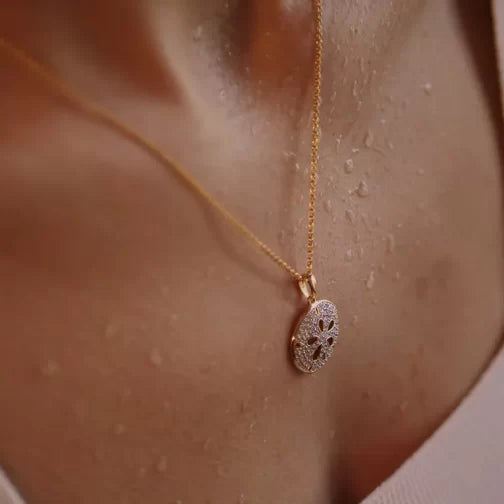 The Gold Vermeil Sand Dollar Necklace by Shanore graces the neck with an ornate pendant, resembling a delicate sand dollar, and complements the slightly glistening skin against a light top.