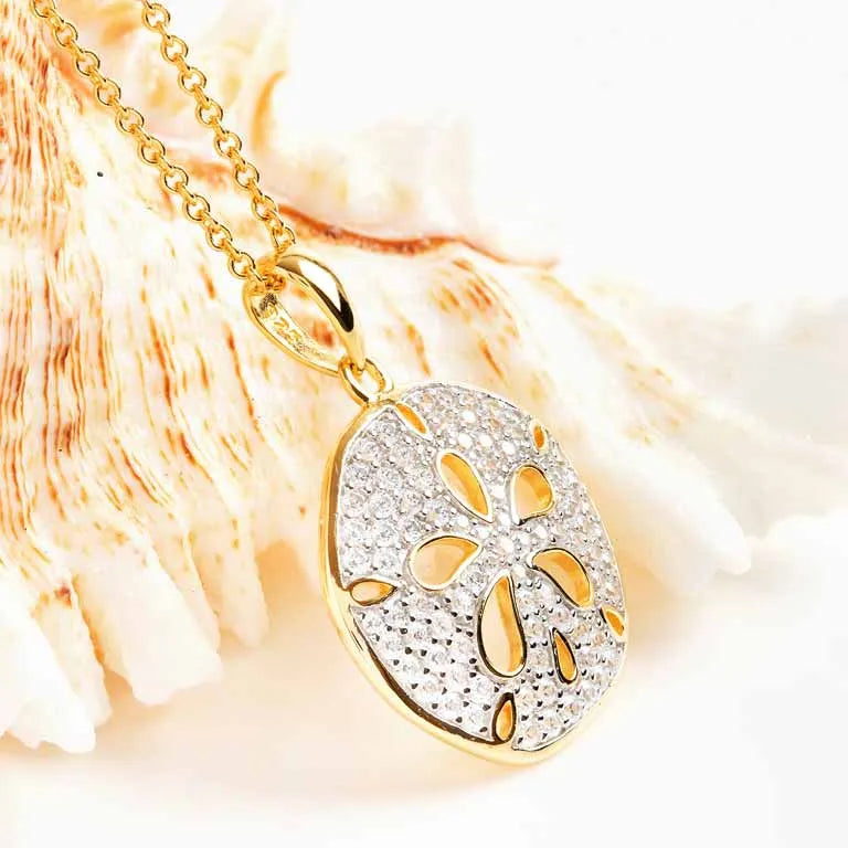 The Shanore Gold Vermeil Sand Dollar Necklace, adorned with cubic zirconia, is beautifully showcased against a seashell backdrop, highlighting its intricate detailing for an elegant beach-inspired look.
