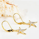 Close-up of Shanores Gold Vermeil Starfish Earrings- Ocean Jewelry, featuring cubic zirconia accents, placed on a light surface beside a textured seashell.