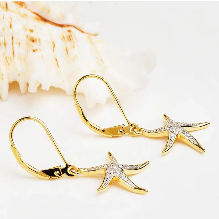 Close-up of Shanores Gold Vermeil Starfish Earrings- Ocean Jewelry, featuring cubic zirconia accents, placed on a light surface beside a textured seashell.