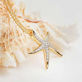 The Shanore Gold Vermeil Starfish Necklace from the Ocean Jewelry collection features a 14k gold vermeil starfish pendant adorned with cubic zirconia stones on a delicate gold chain. A light-colored seashell in the softly lit background enhances its elegance.