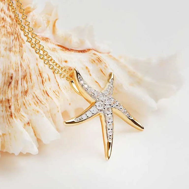 The Shanore Gold Vermeil Starfish Necklace from the Ocean Jewelry collection features a 14k gold vermeil starfish pendant adorned with cubic zirconia stones on a delicate gold chain. A light-colored seashell in the softly lit background enhances its elegance.