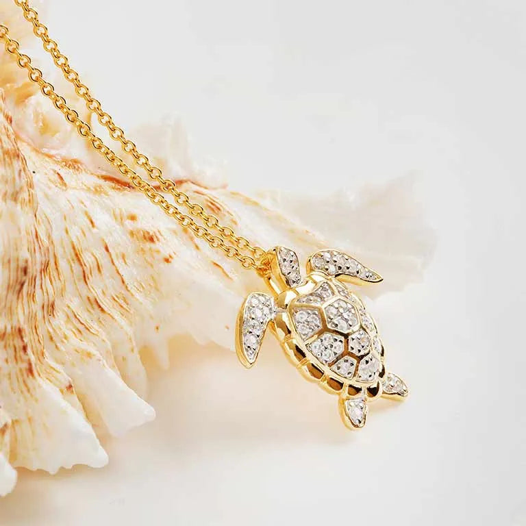 The Shanore Gold Vermeil Turtle Necklace, from the Ocean Jewelry collection, features a turtle-shaped pendant crafted in 14k gold with sparkling cubic zirconia on a gold chain, set against a textured seashell backdrop.