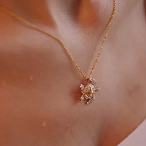 A close-up shows a woman wearing the Gold Vermeil Shell Turtle Necklace by Shanore, featuring a delicate pendant encrusted with sparkling cubic zirconia. It hangs elegantly against her skin while a softly blurred background enhances its charm.