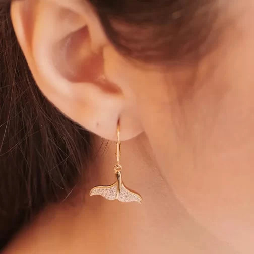 A person’s ear is adorned with Shanores Gold Vermeil Whale Tail Drop Earrings, part of their Ocean Jewelry collection. Elegantly embellished with sparkling cubic zirconia stones, the earrings are beautifully highlighted against a softly blurred background.