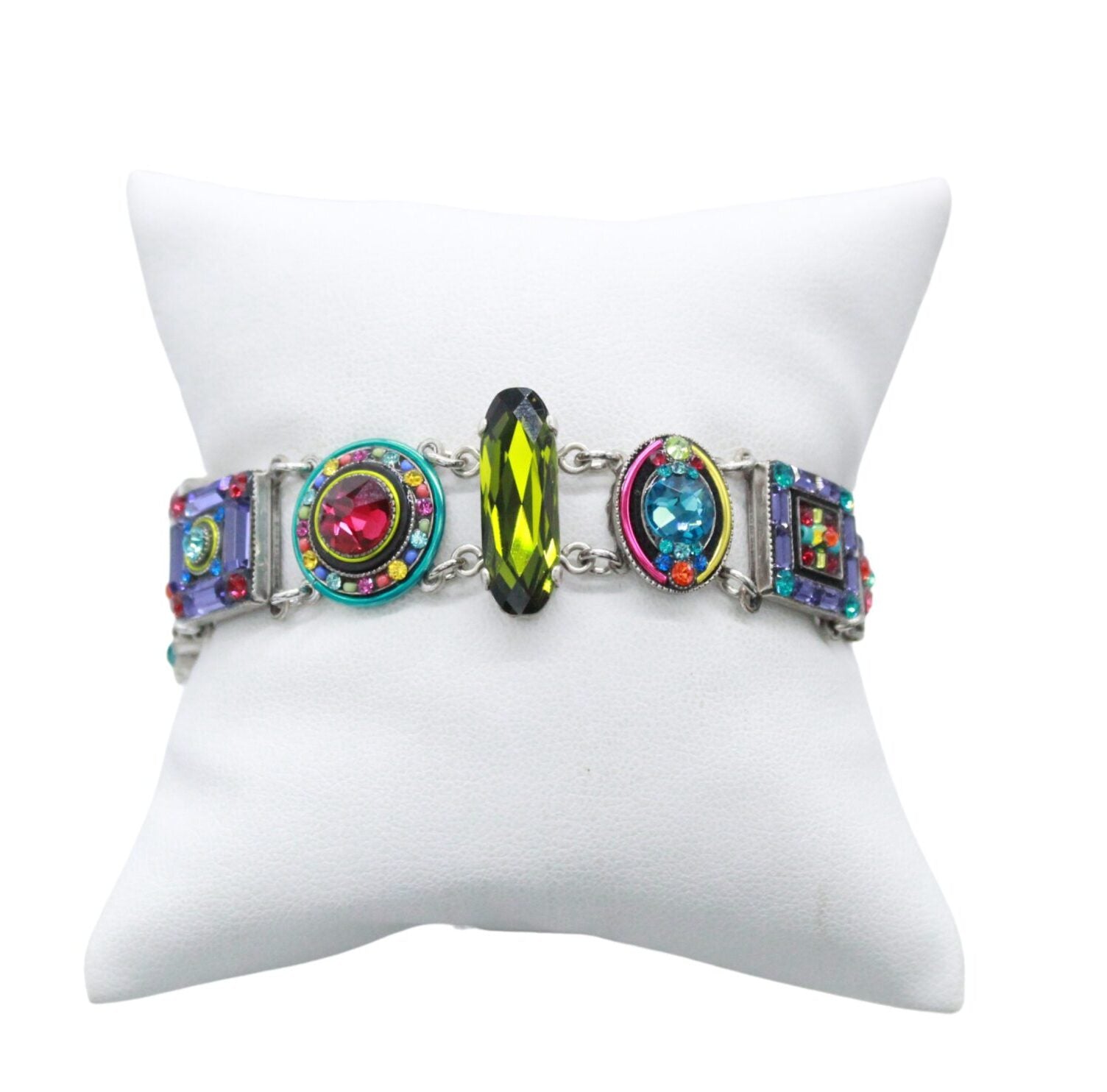 The La Dolce Vita Multi-Color Bracelet by Firefly, a handmade piece, is displayed on a white cushioned stand. Geometric shapes with green, pink, and blue gemstones contrast strikingly while honoring fair-trade practices.