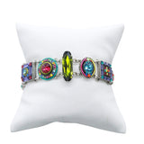 The La Dolce Vita Multi-Color Bracelet by Firefly, a handmade piece, is displayed on a white cushioned stand. Geometric shapes with green, pink, and blue gemstones contrast strikingly while honoring fair-trade practices.
