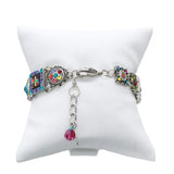 Presenting the Firefly La Dolce Vita Multi-Color Bracelet, a handmade jewel with intricate patterns and multicolored accents on a white cushion backdrop. It includes square and round elements, a chain clasp, and features a dangling red bead charm. Crafted using fair-trade practices.